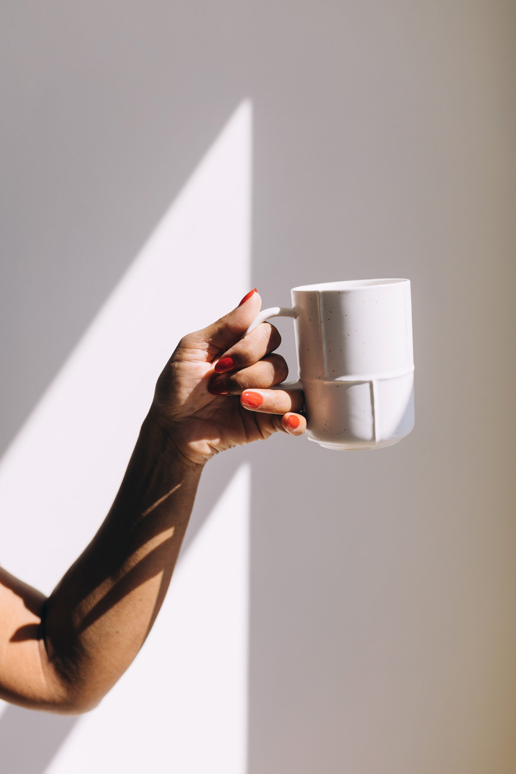 Woman with orange nail polish holding up coffee mug against a plain wall with light filtering through nearby window.